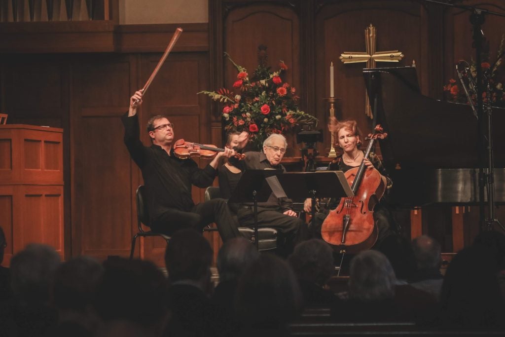 A violinist, cellist, and pianist performing with bows and hands in the air.
